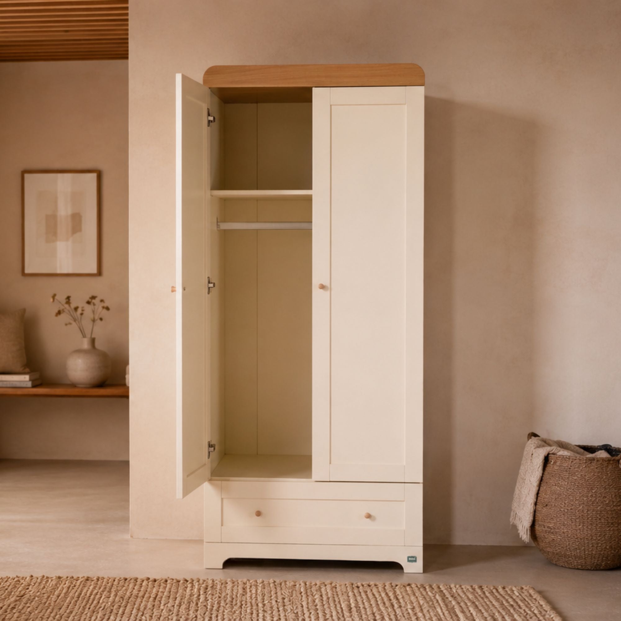 A tall cream-colored wooden wardrobe with one door open, showing an interior shelf and hanging rod, set in a warm, minimalist room with a woven basket and neutral wall.