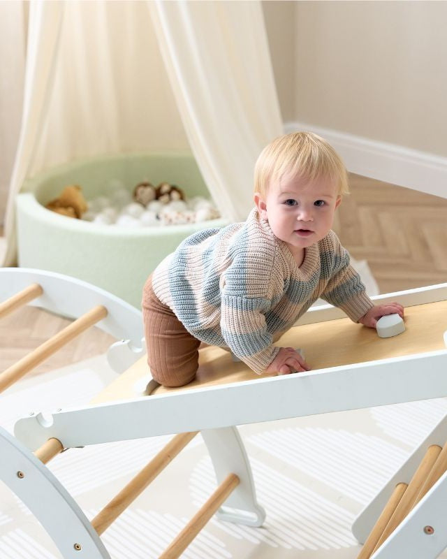 Toddler standing on a wooden climbing toy with a ball pit in the background