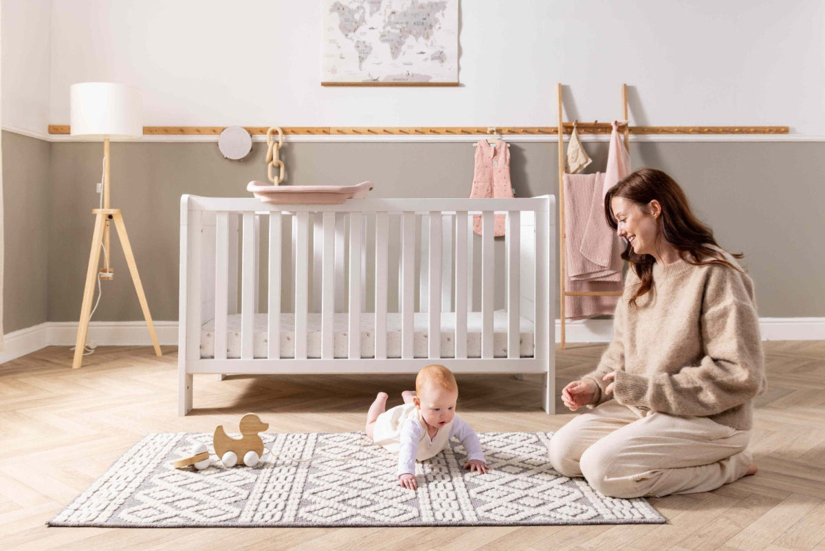A woman with a baby in a nursery with a white cot bed, Cushi cot top changer and a grey and white rug.