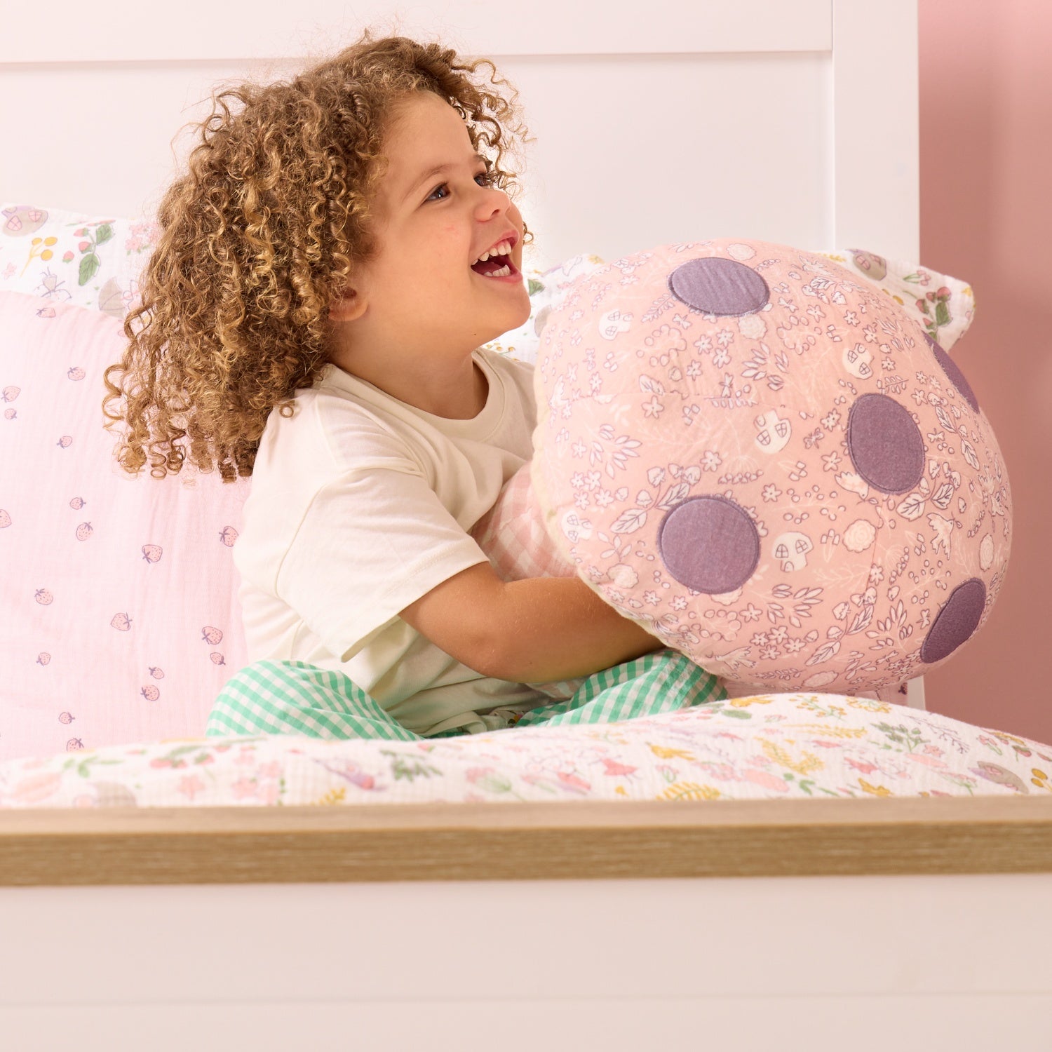Smiling child holding the Soft Muslin Mushroom Cushion on a bed, showing its playful spotted top and soft pastel fabric textures