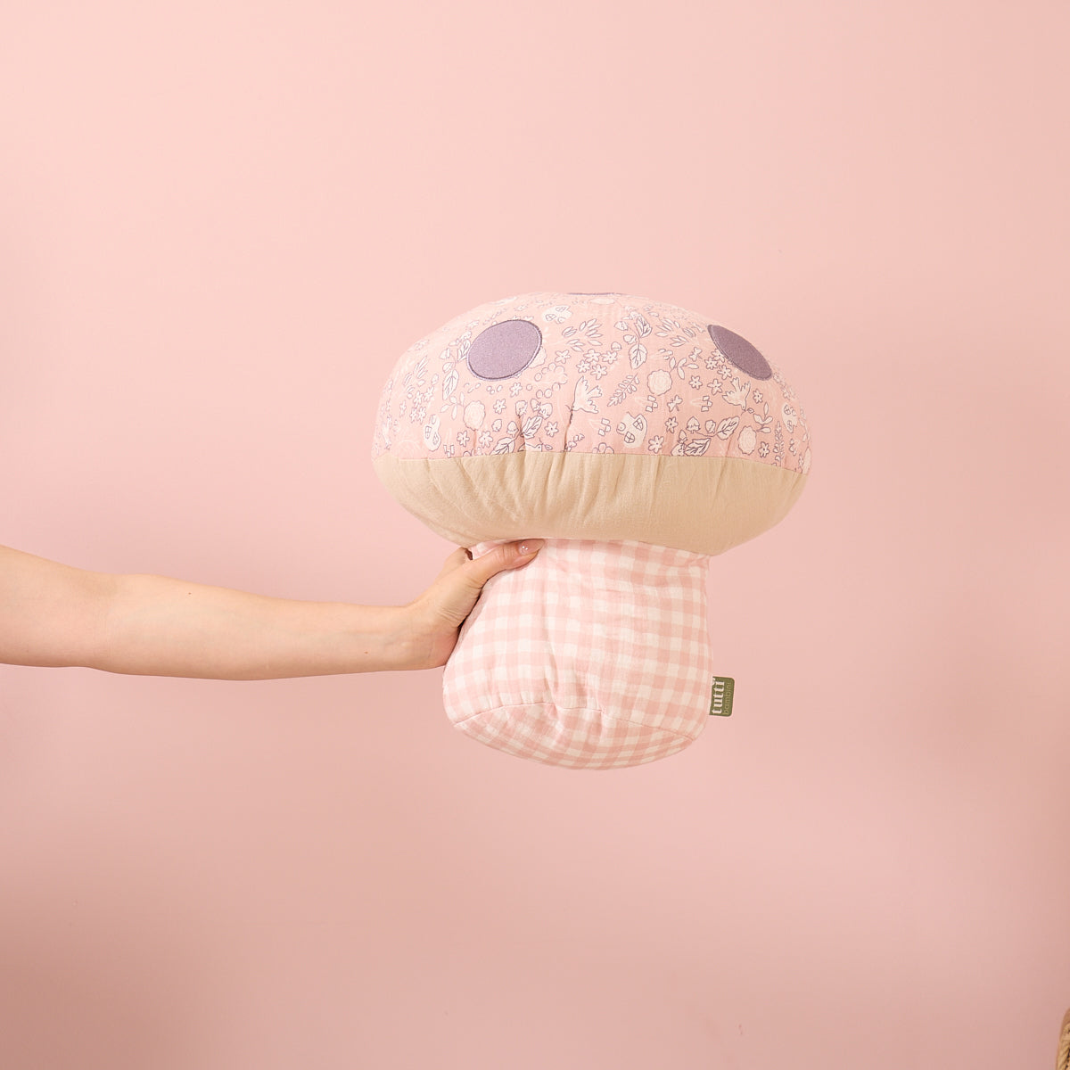 Hand holding the Soft Muslin Mushroom Cushion against a pink background, highlighting its rounded shape and gingham stem detail