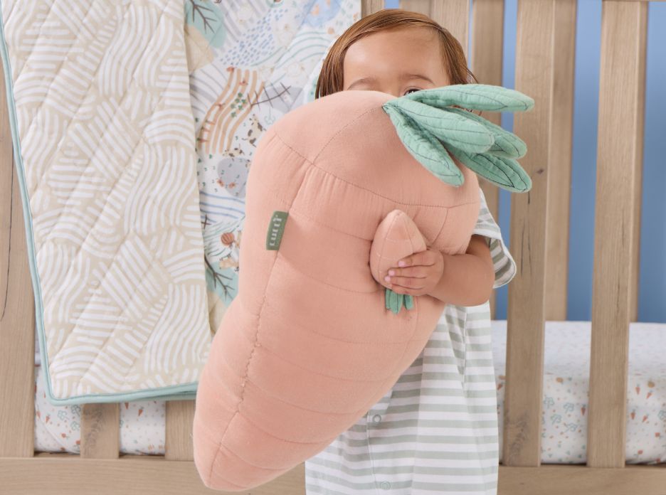 lifestyle of toddler cuddling oversized soft muslin carrot cushion in a cot styled with woodland bedding in calming pastel tones.