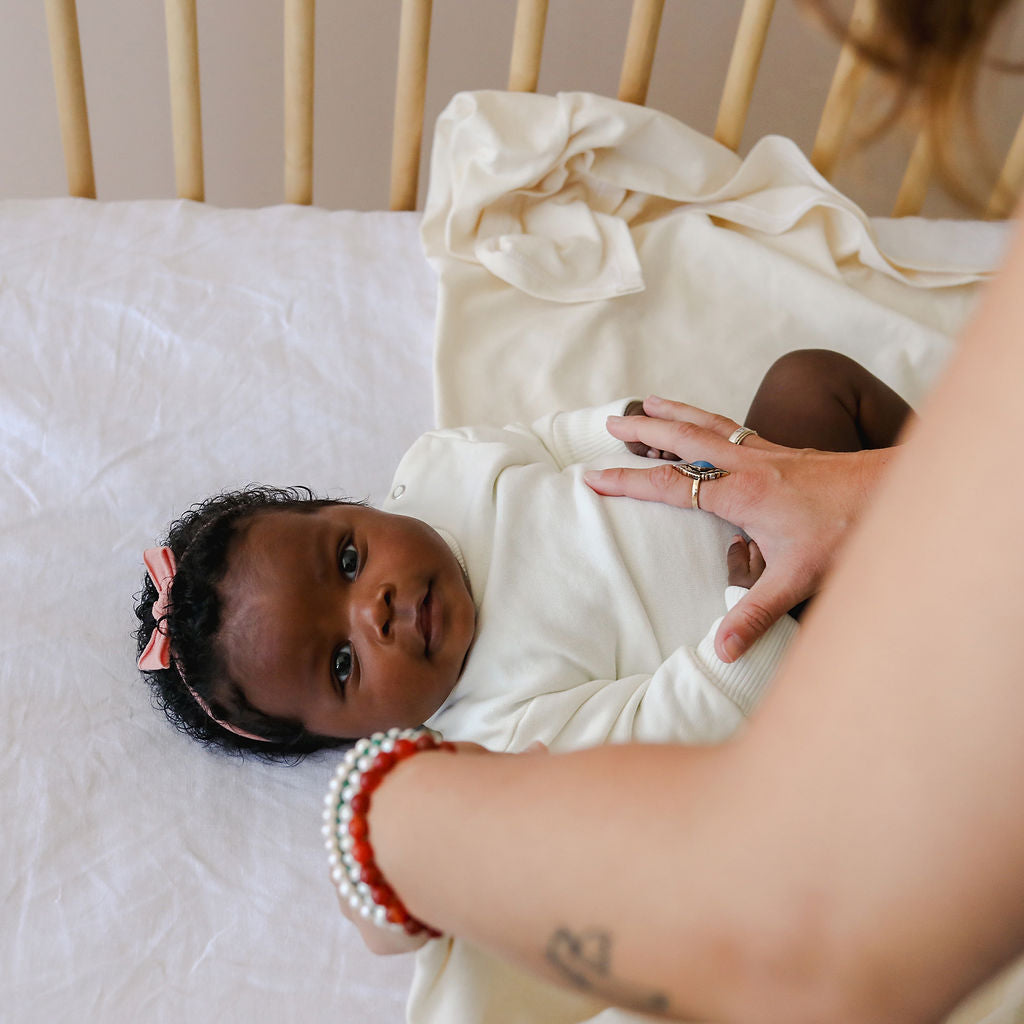 Baby wrapped in a white blanket with a person&#39;s hand holding it, lying on a white surface.