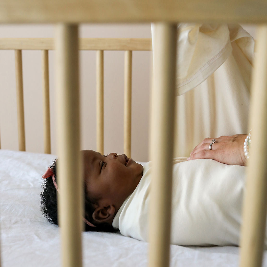 Baby lying in a crib with a person's hand gently touching the crib rail.