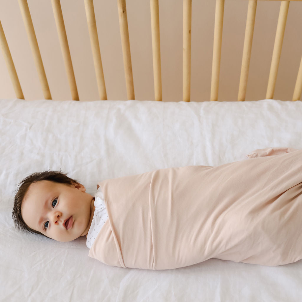 Newborn baby swaddled in a light pink blanket on a white crib with wooden slats.