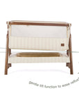 An infographic of a baby bedside crib with stiped cream-colored fabric panels and a matching base, shown on a white background with a dashed horizontal line indicating the tilt function and text reading 'gentle tilt function to ease reflux'.