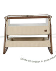 An infographic of a baby bedside crib with cream quilted fabric panels and a mesh upper screen, shown on a white background with overlaid text and arrows explaining the gentle tilt function to ease reflux.