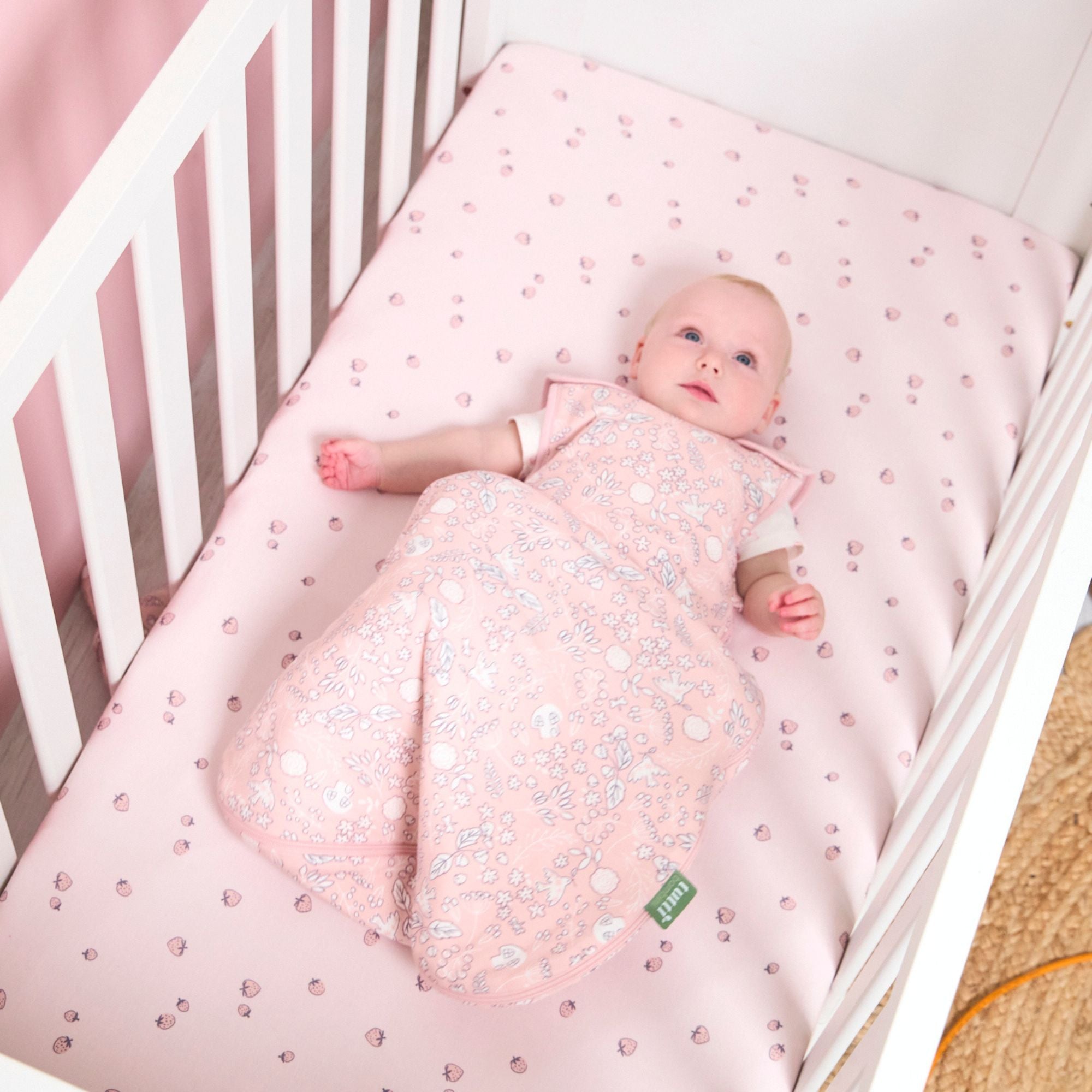 Baby lying in a cot bed wearing a sleep bag, on pink printed fitted sheets.