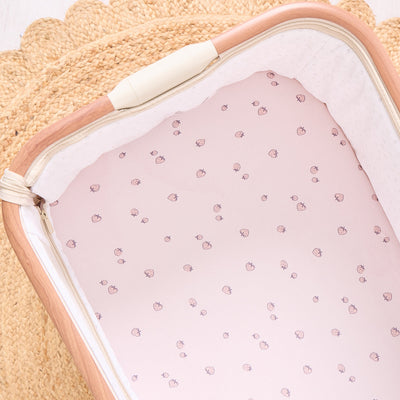 Close-up of strawberry print tiny tails  bedside crib fitted sheet with wooden frame and woven storage basket backdrop.