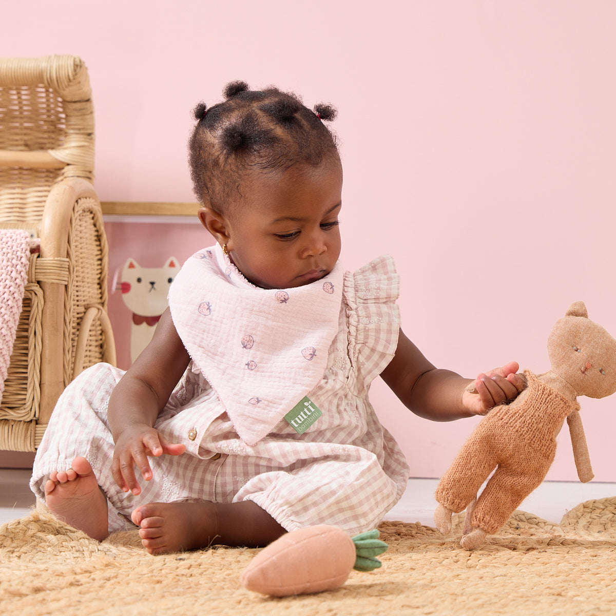 Baby wearing the Tiny Tails bandana, playing with a toy on a soft surface with a pink background
