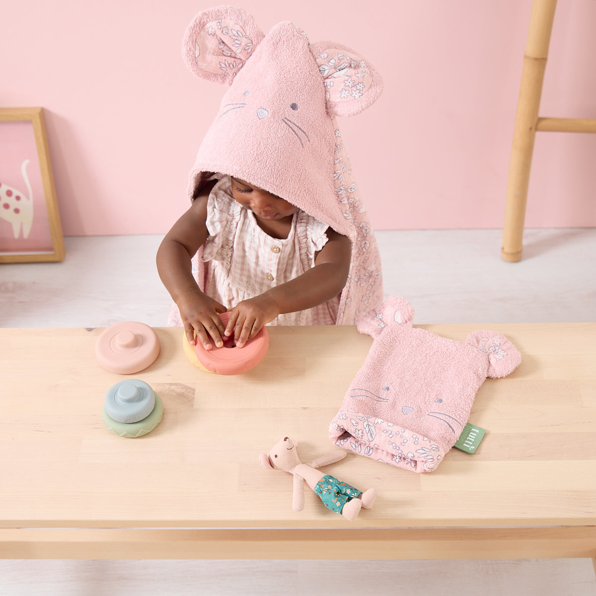 Baby wearing Tiny Tails pink hooded towel with embroidered mouse face and floral print lining, styled during playtime.