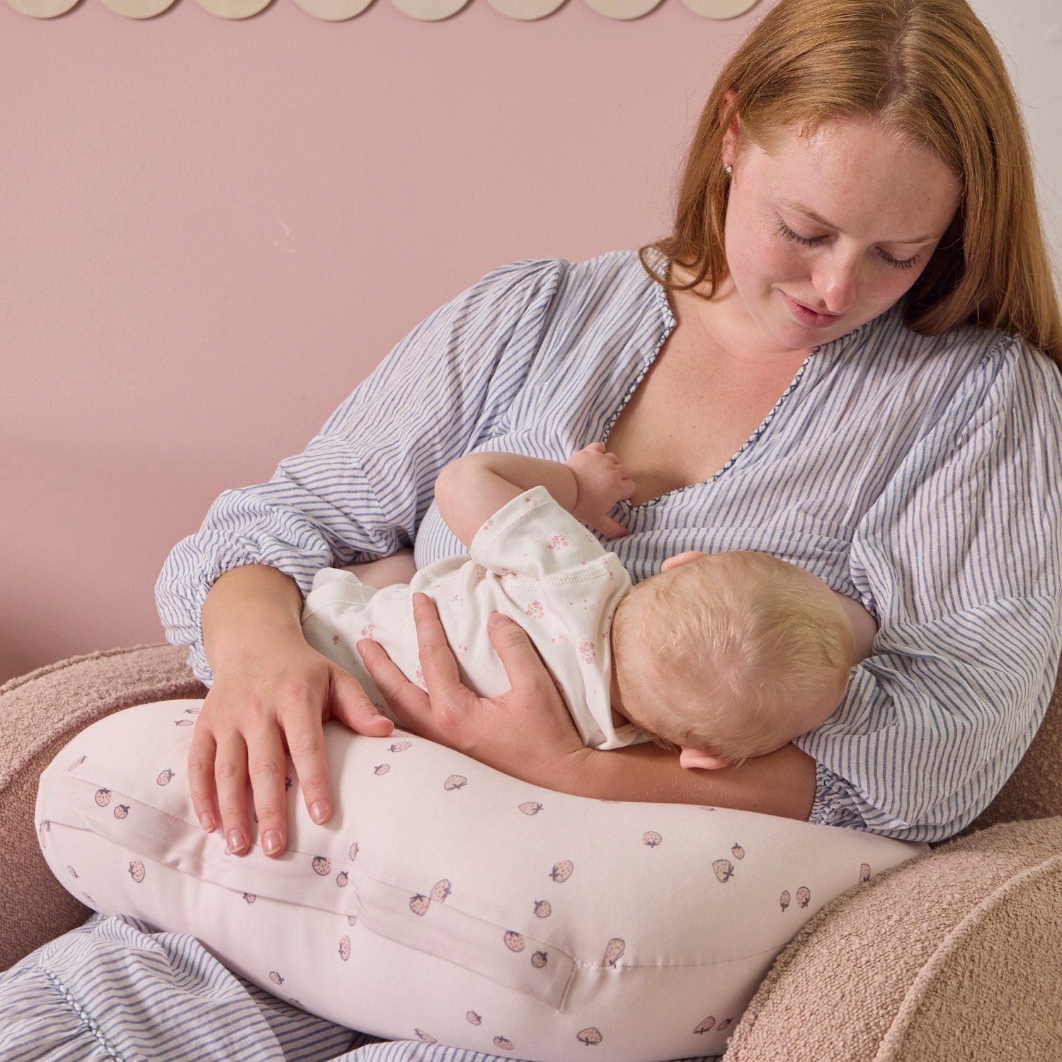 A woman breastfeeding a baby using the Tiny Tails nursing pillow in pink with a strawberry pattern, against a pink wall.