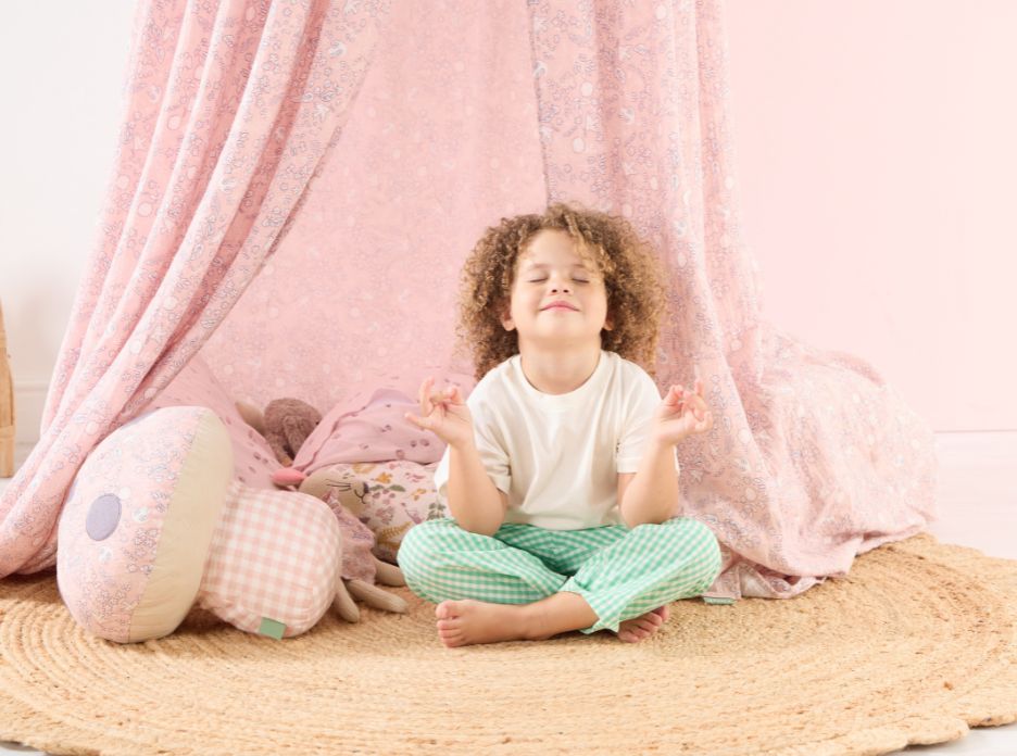 Child sitting cross-legged beneath the Tiny Tails canopy, creating a calm reading nook with soft pink fabrics and cushions.