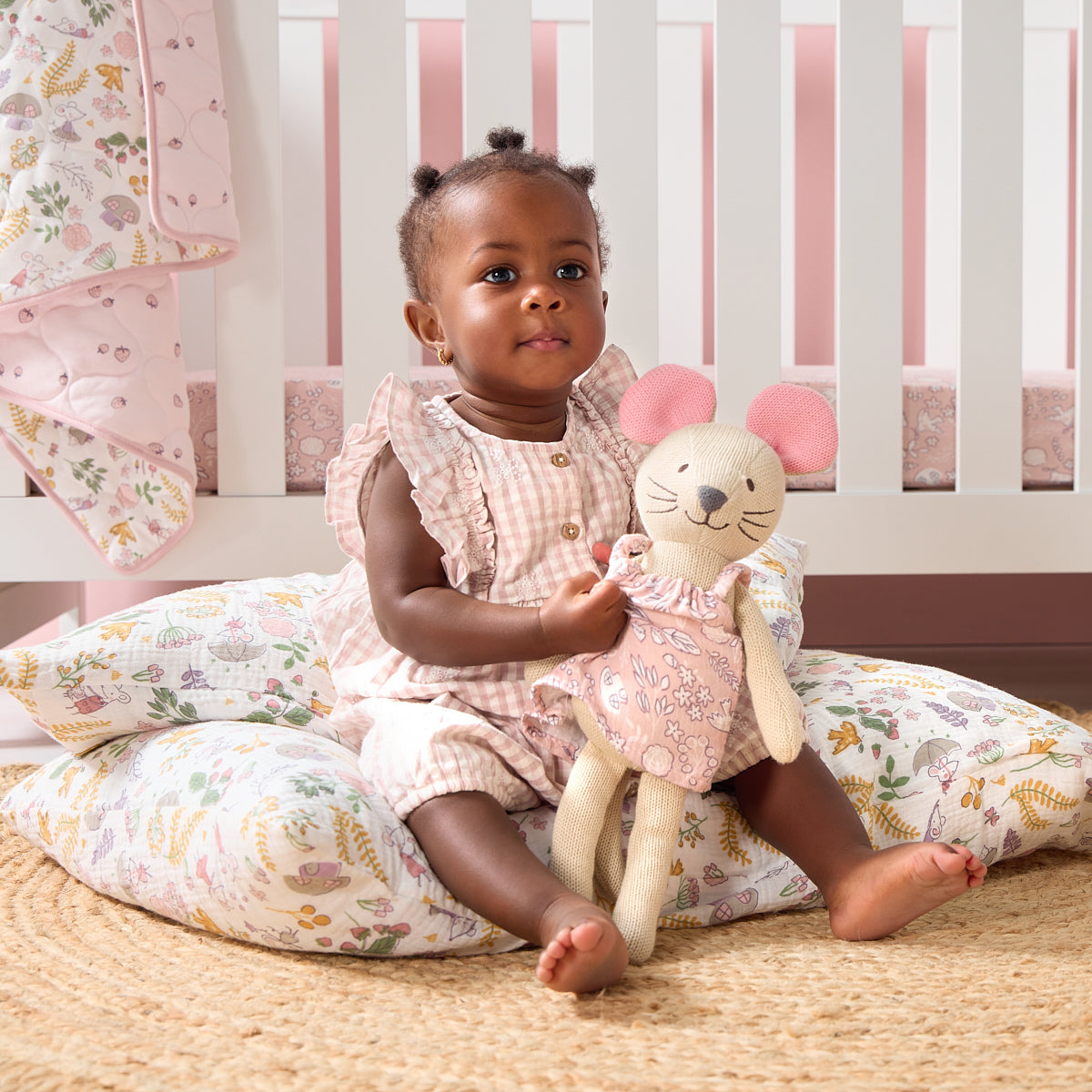 Child sitting on a floral pillow in a cot, holding a stuffed mouse teddy in a cosy nursery setting.