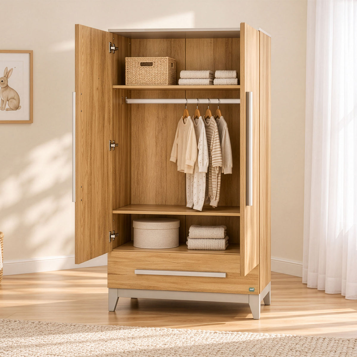 A golden oak wood two-door wardrobe with handles and legs, shown open with baby clothes and storage baskets inside, set in a brightly lit room with a wooden floor and white curtains.