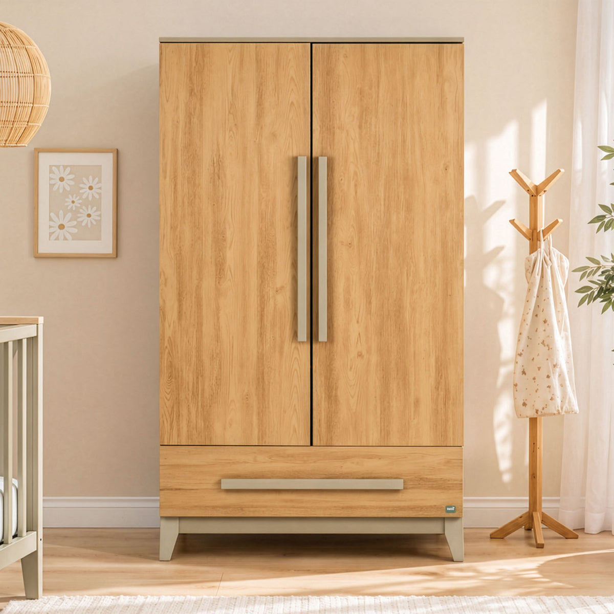 A tall wooden two-door nursery wardrobe with a lower drawer, shown in a styled nursery or children's room. The wardrobe has a golden oak wood grain finish with sage handles and a sage green base.