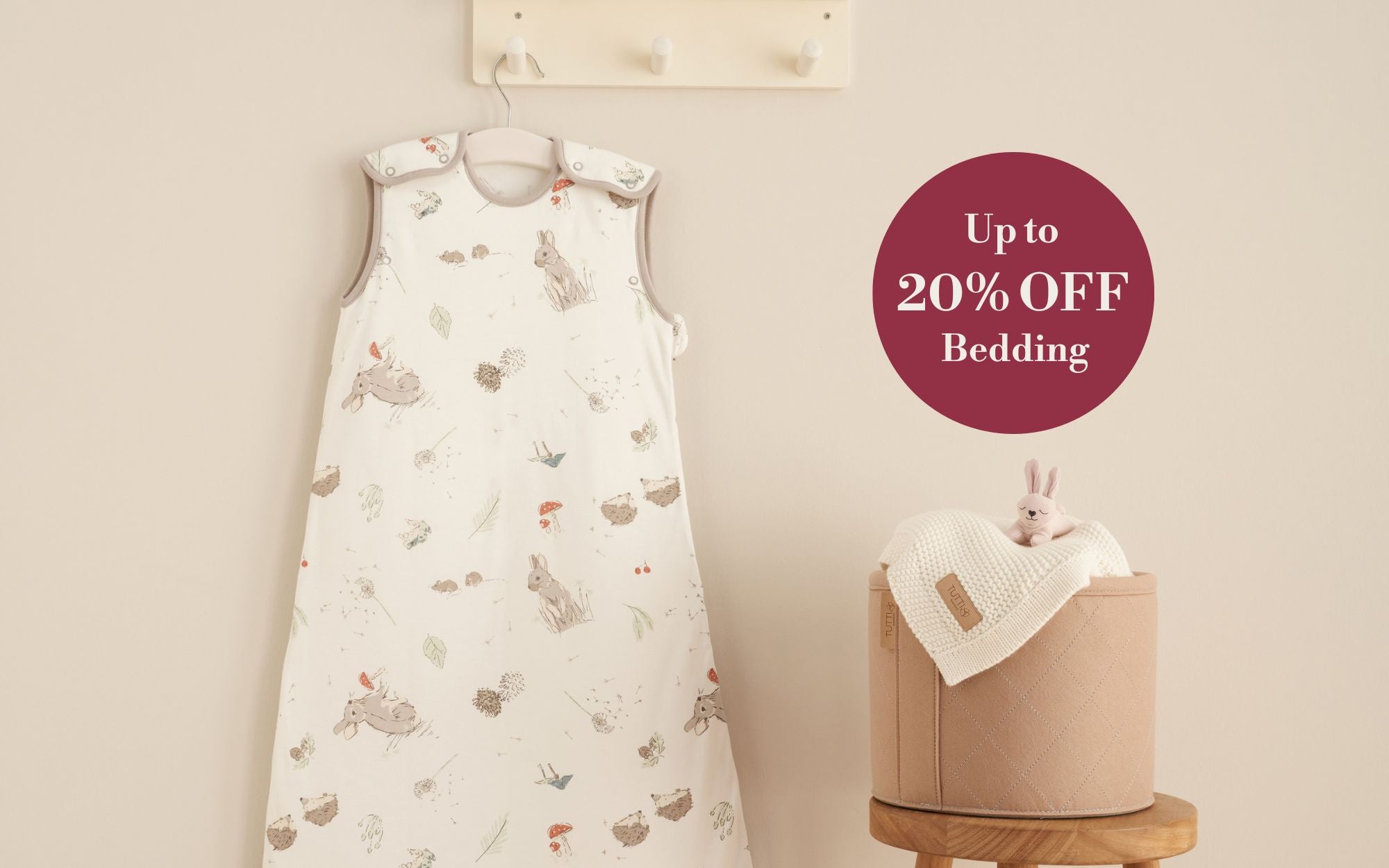 Baby slep bag with animal patterns on a hanger, next to a basket with a blanket and toy, against a beige wall with a discount label.