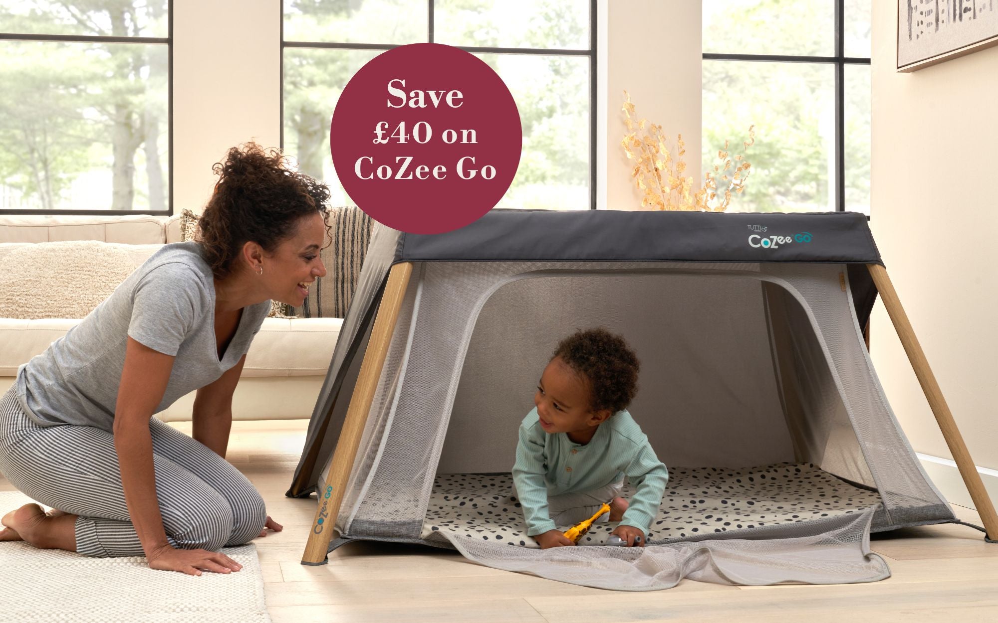 Woman and child playing in a CoZee Go travel cot in a living room.