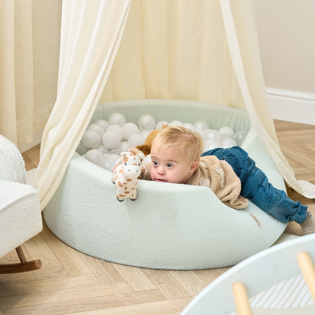 Baby lying comfortably over the padded edge of the Bola baby ball pit in sea mist, enjoying soft textures and sensory playtime