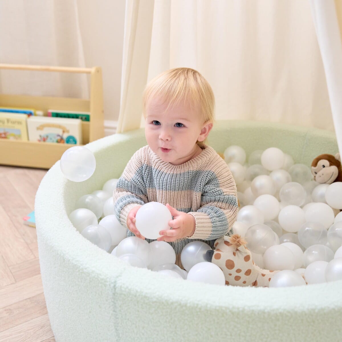 Toddler sitting inside the Bola baby ball pit in sea mist, holding play balls to encourage tactile exploration and early motor skills