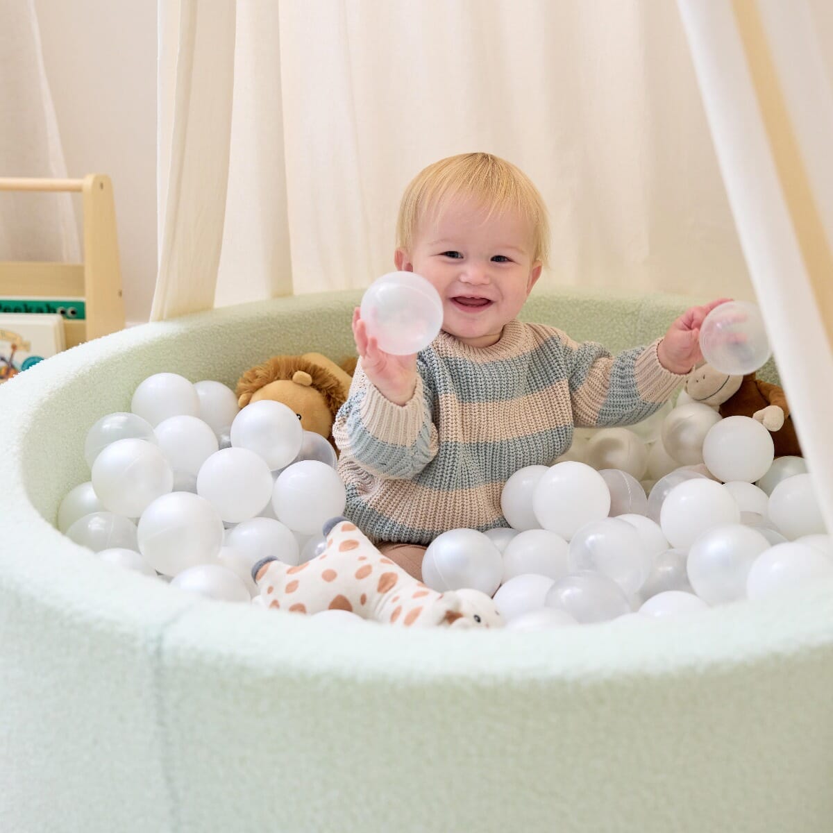 Smiling toddler playing inside the Bola baby ball pit in sea mist, holding soft balls during relaxed sensory playtime at home