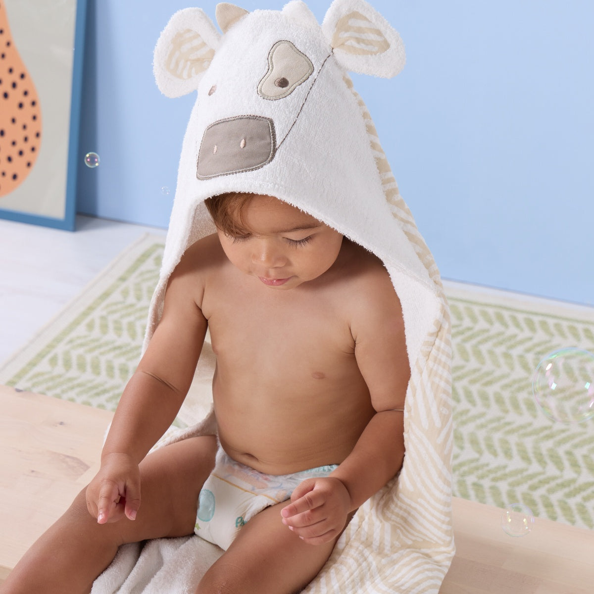 Toddler wrapped in Clemi the Cow hooded towel, hood up to show embroidered cow face, soft ears, and patterned lining
