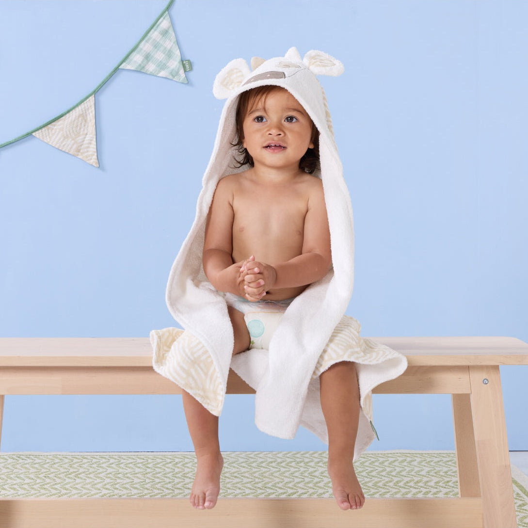 Toddler sitting on a bench wrapped in the Clemi the Cow hooded towel, featuring soft beige horns and patterned inner lining in gentle tones