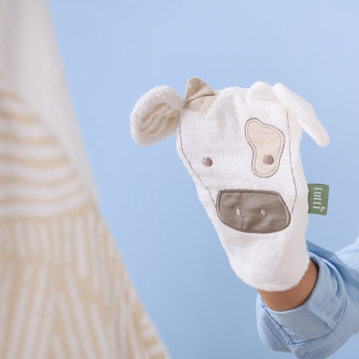 Close-up of Clemi the Cow wash mitt in soft white terry fabric with embroidered cow face, gentle beige accents, and Tutti Bambini tag visible