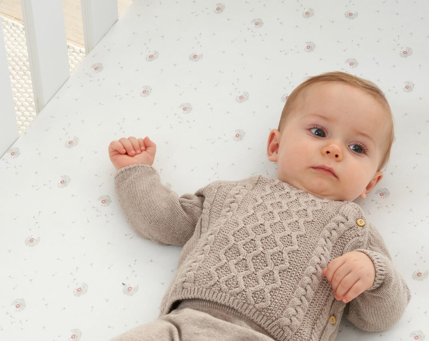 Baby lying on Cocoon print cot bed fitted sheet with soft dandelion pattern, highlighting breathable cotton fabric and gentle, cosy design