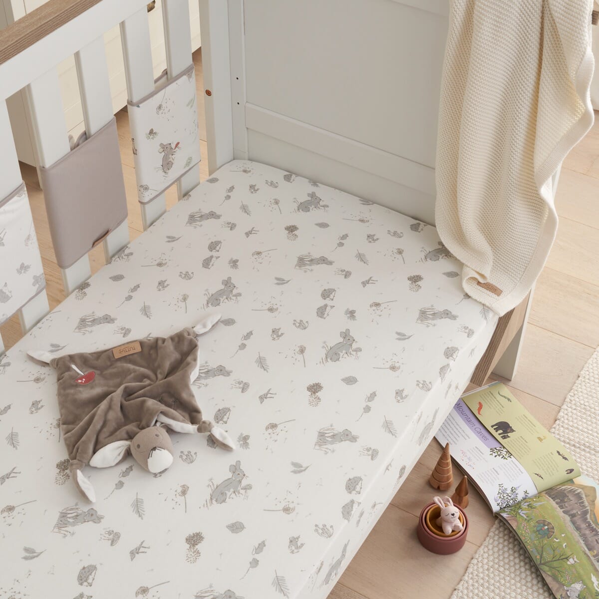 Lifestyle nursery view of Cocoon cot bed fitted sheet with woodland rabbit design, styled with soft blanket and toys for a cosy bedtime setting