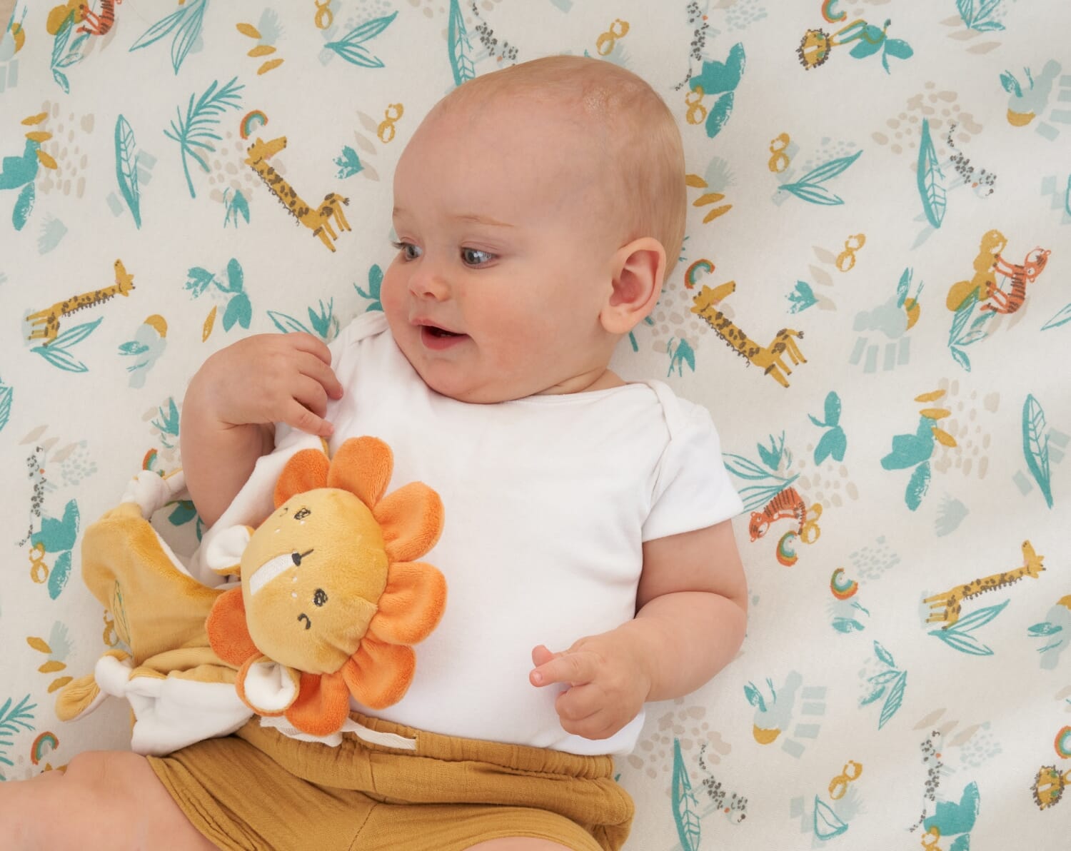 Baby lying on Run Wild cot bed fitted sheet with playful safari print, holding a soft lion comforter on a cosy, patterned sleep surface