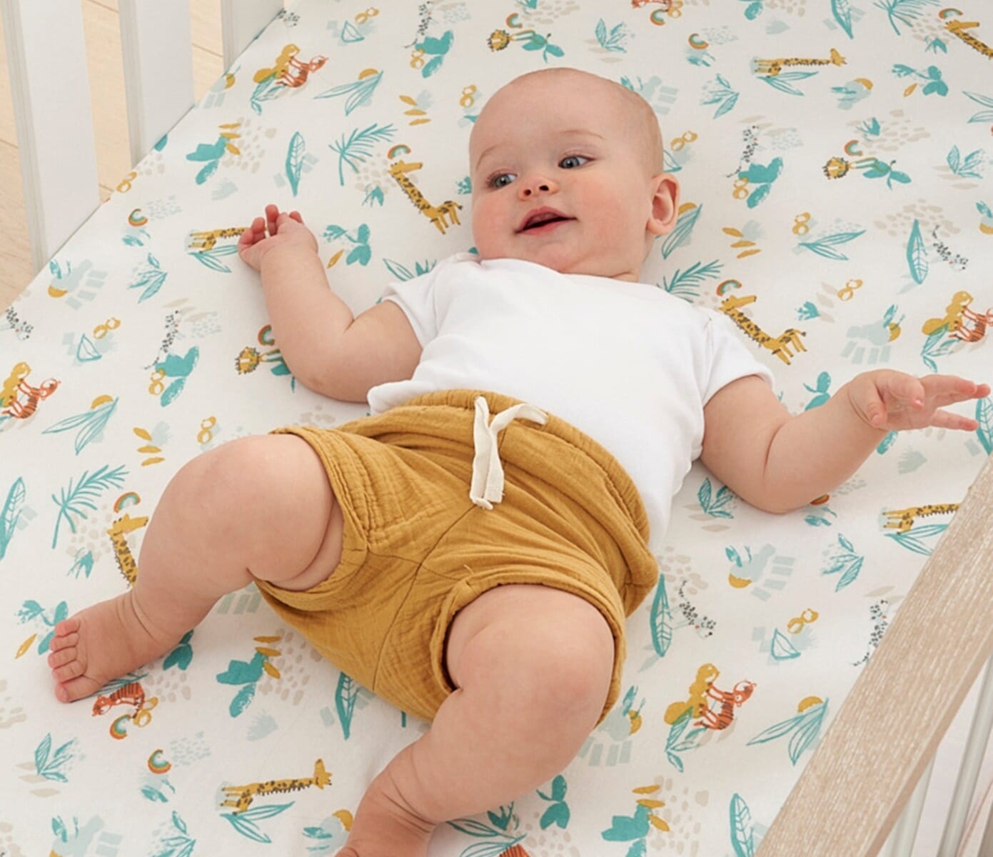 Smiling baby lying on Run Wild cot bed fitted sheet with vibrant safari illustrations, adding colour and comfort to their cosy sleep space