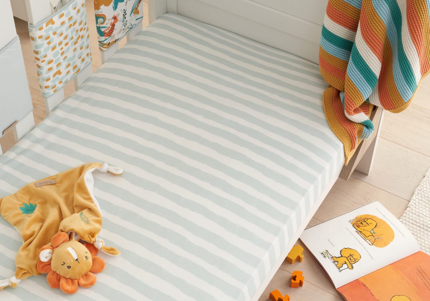 Striped Run Wild cot bed sheet styled with a lion comforter and open storybook, creating a warm, inviting nursery play and rest space