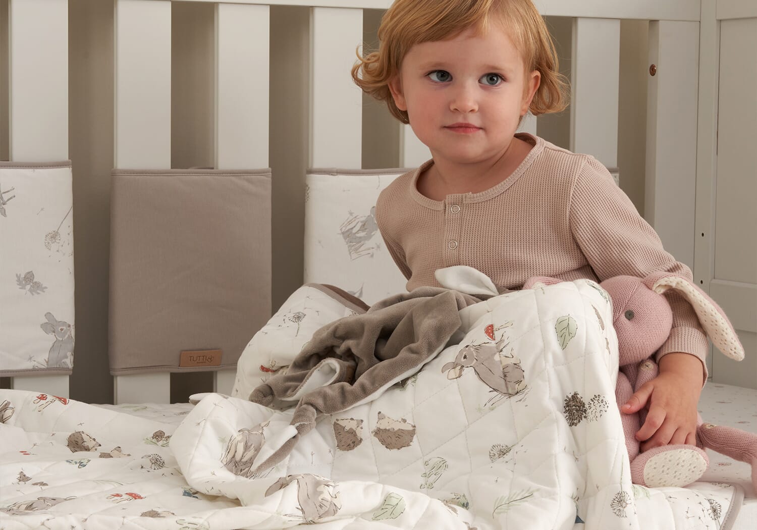 Toddler sitting up on a cot bed with the Cocoon coverlet, showcasing the soft quilted design and gentle woodland print in a calm nursery setting