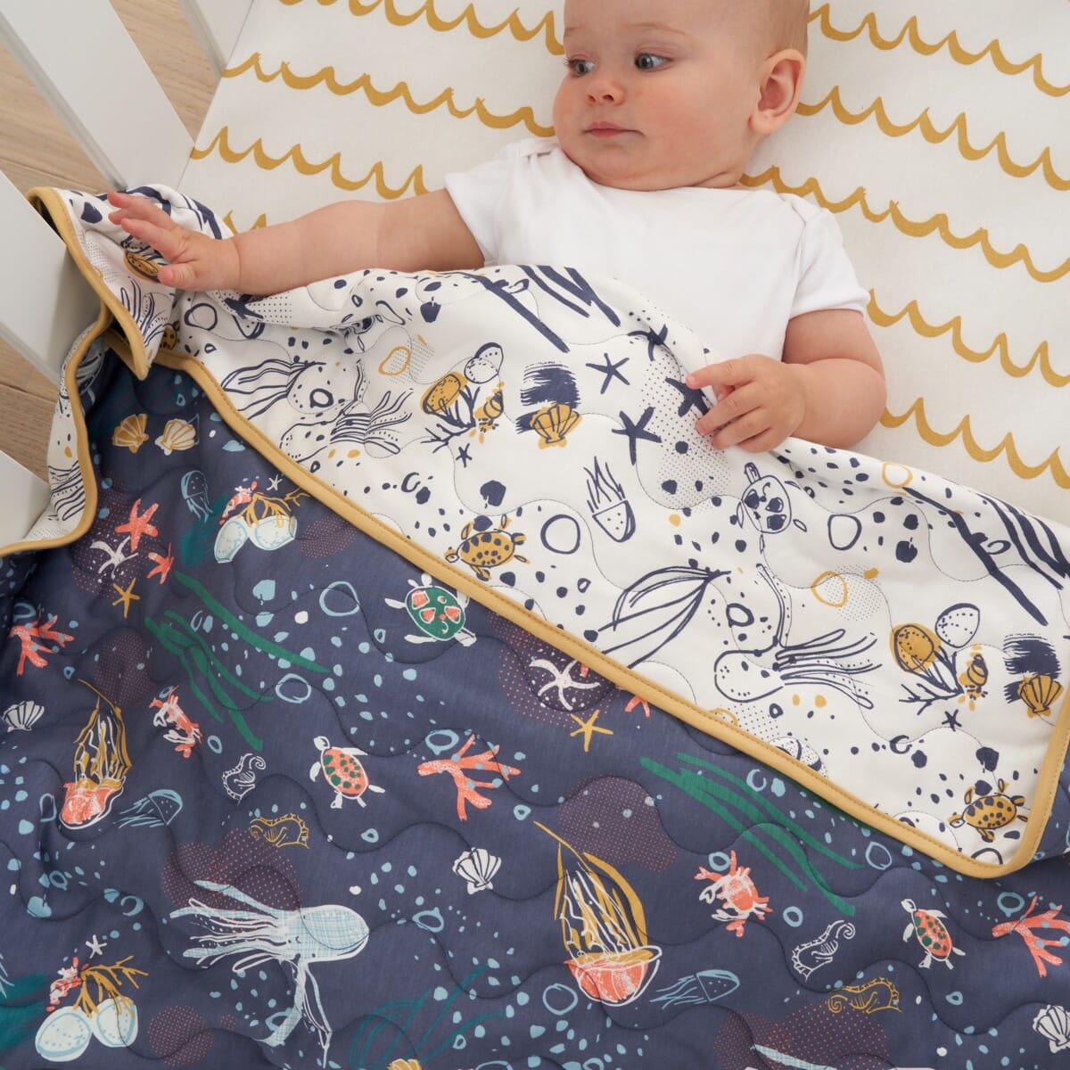 Baby tucked under Our Planet coverlet, reversible quilted cotton with ocean-themed prints and mustard trim for warmth and playful comfort