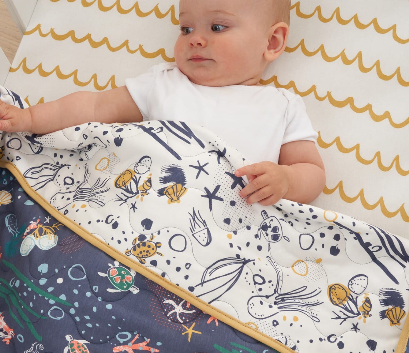 Feature image of baby tucked under Our Planet coverlet, reversible quilted cotton with ocean-themed prints