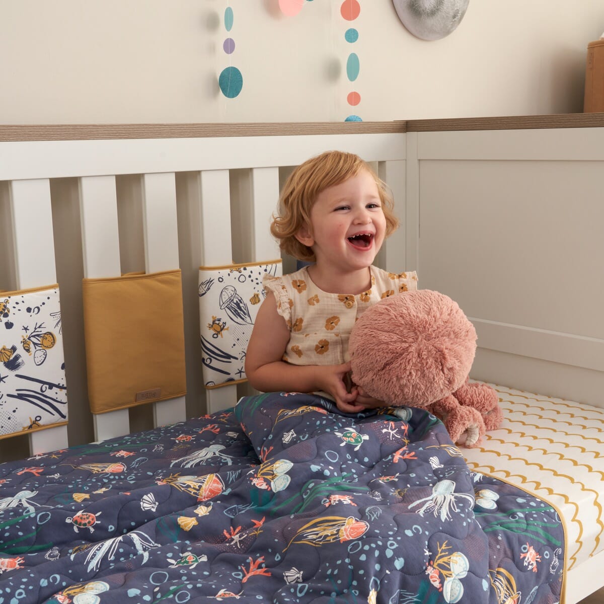 Smiling toddler with Our Planet coverlet, quilted cotton featuring ocean-inspired prints offering warmth, comfort, and playful charm