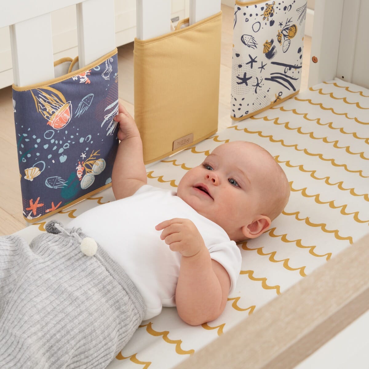 A baby lying on a cot with a yellow and white striped cot wrap around the cot. The wrap features sea life motifs with colorful fish and coral.