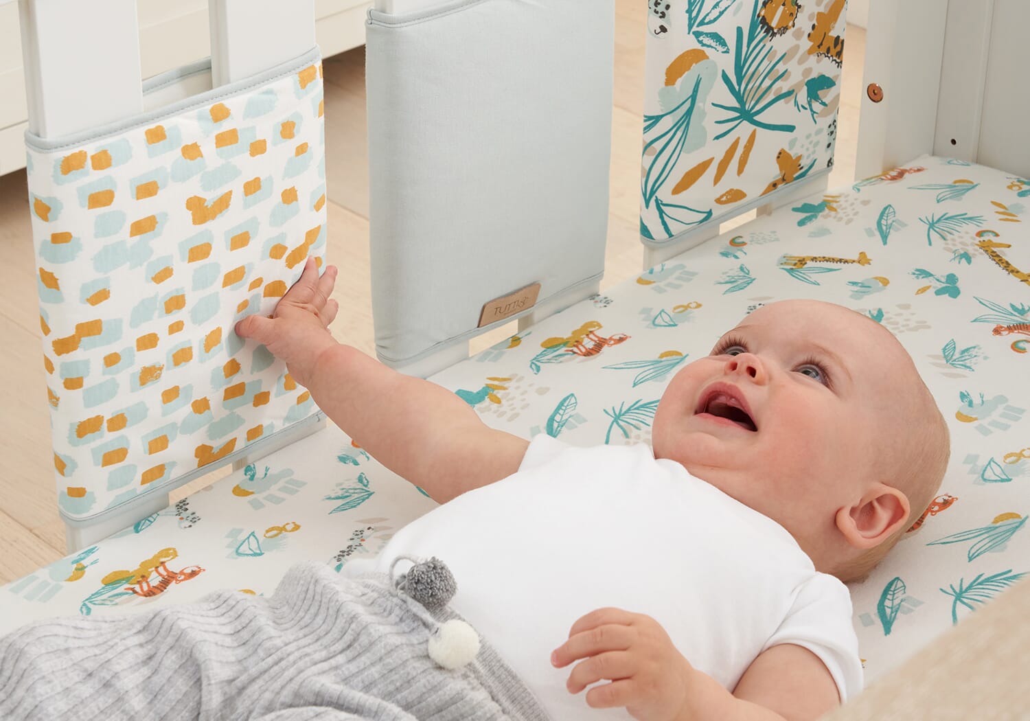 Baby reaching towards Run Wild cot wraps, showing padded protection on cot rails with playful prints designed for everyday nursery use