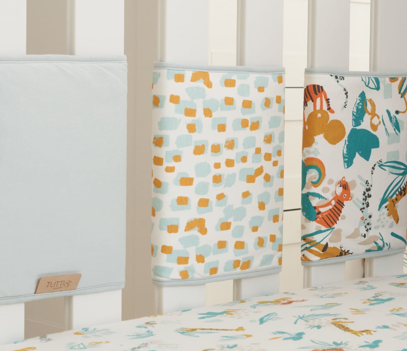 Close-up of Run Wild cot wraps secured to cot rails, showing softly padded panels with coordinated plain and patterned designs for gentle protection