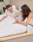 Family relaxing in bed beside the CoZee bedside crib in oak and charcoal, illustrating cosy side-sleeping setup for bonding and reassurance