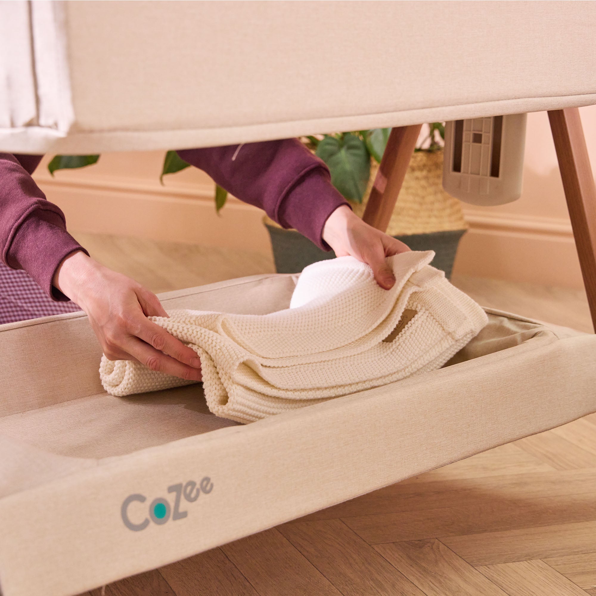 View of CoZee bedside crib in Scandinavian walnut and ecru with parent placing blanket in handy under-crib storage shelf