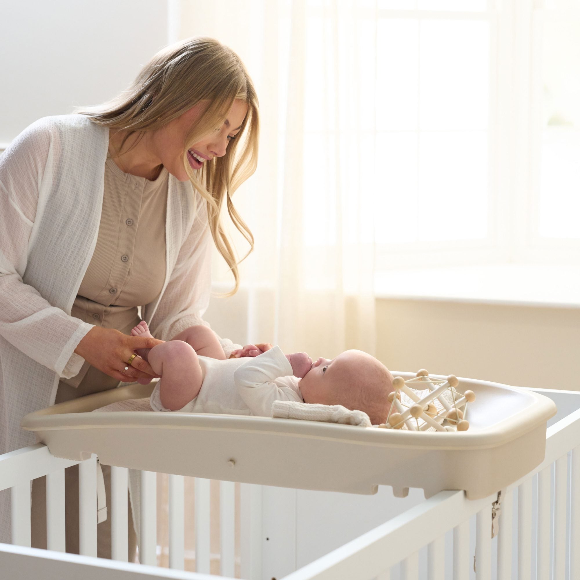 Mum smiling while changing baby on the Cushi Baby Changing Mat in cashmere, showing secure cot-top fit and cosy design