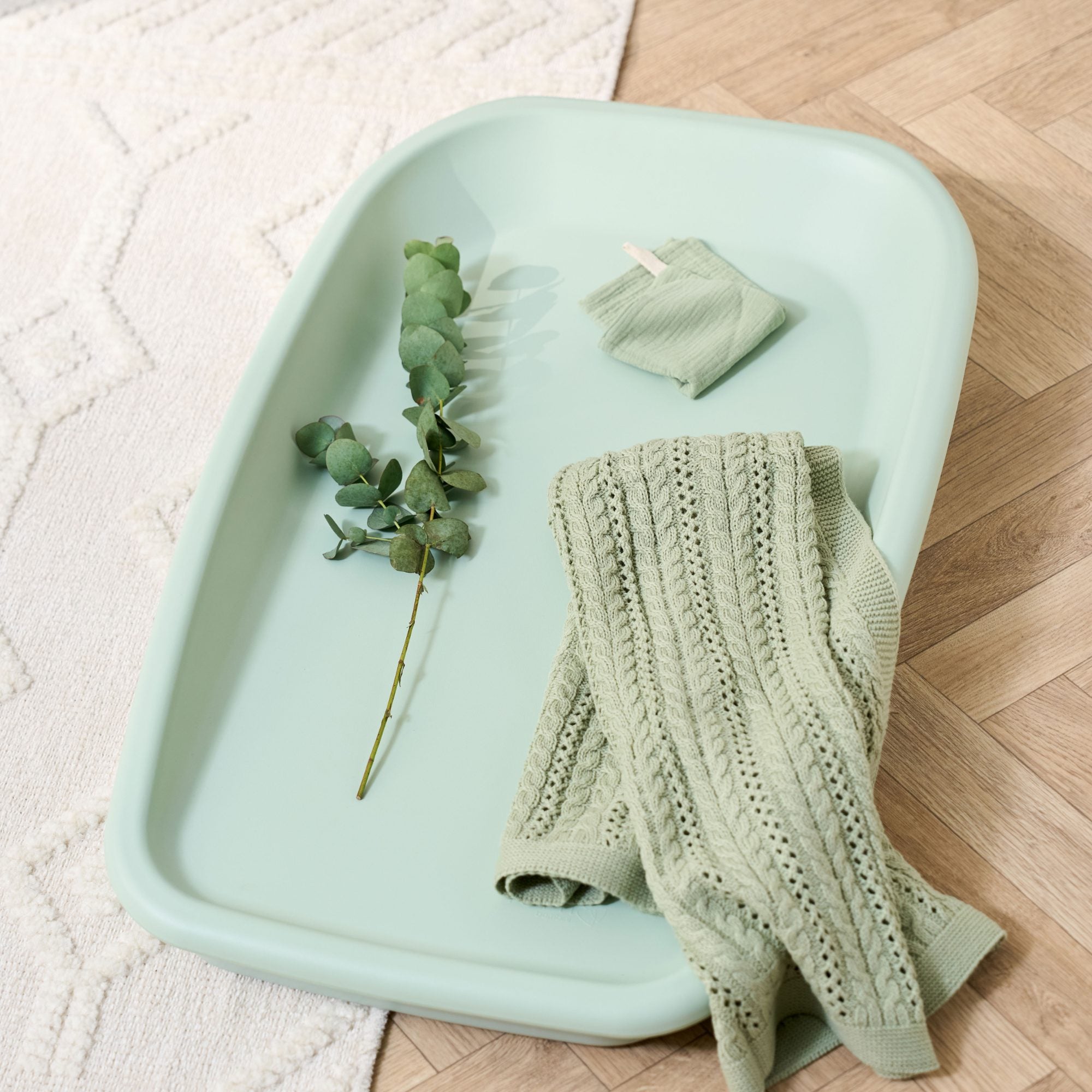 Styled lifestyle view of the Cushi changing mat in leaf green, shown with knitted blanket and eucalyptus for nursery detail