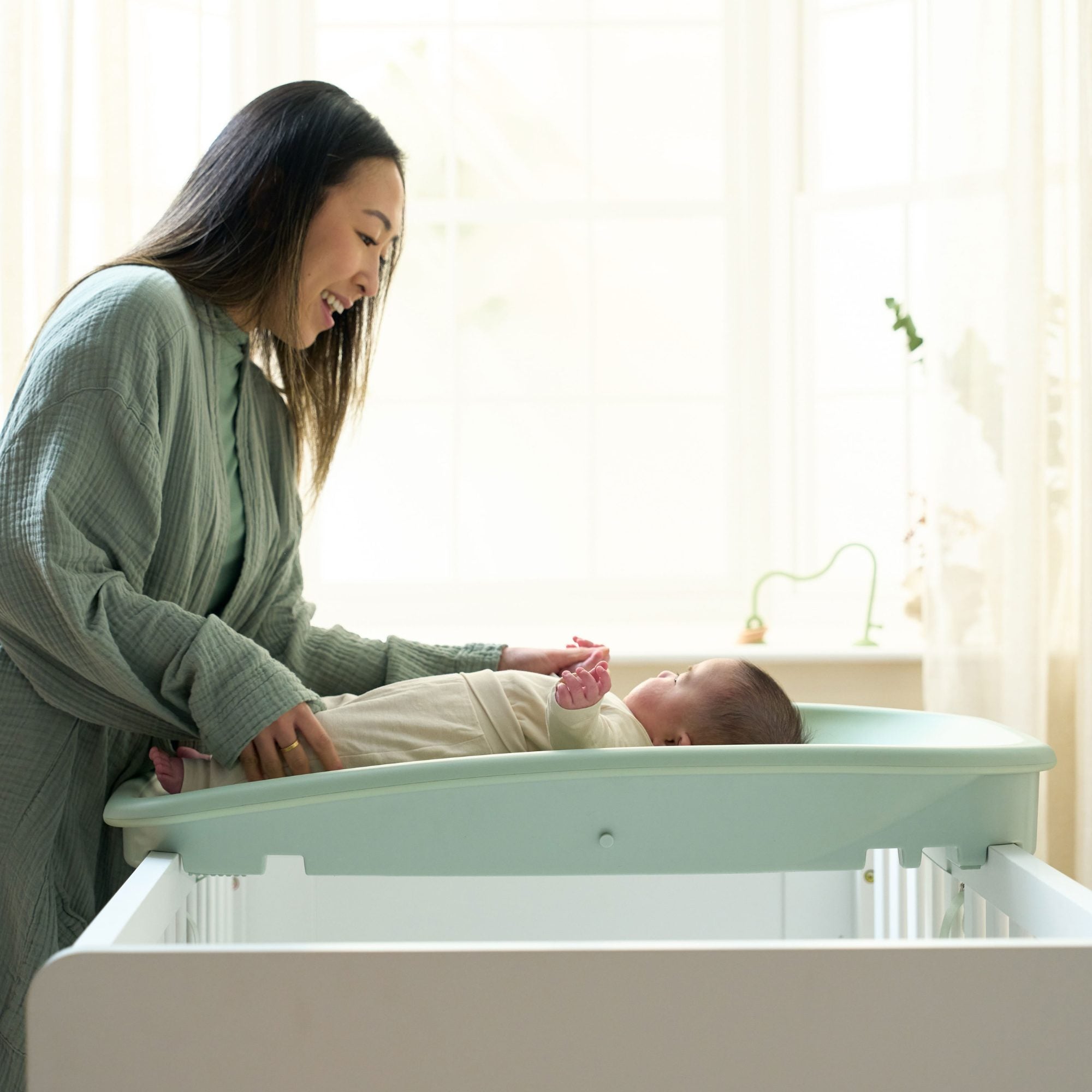 Mother interacting with her baby on the Cushi changing mat in leaf green, fitted securely across the top of a cot bed