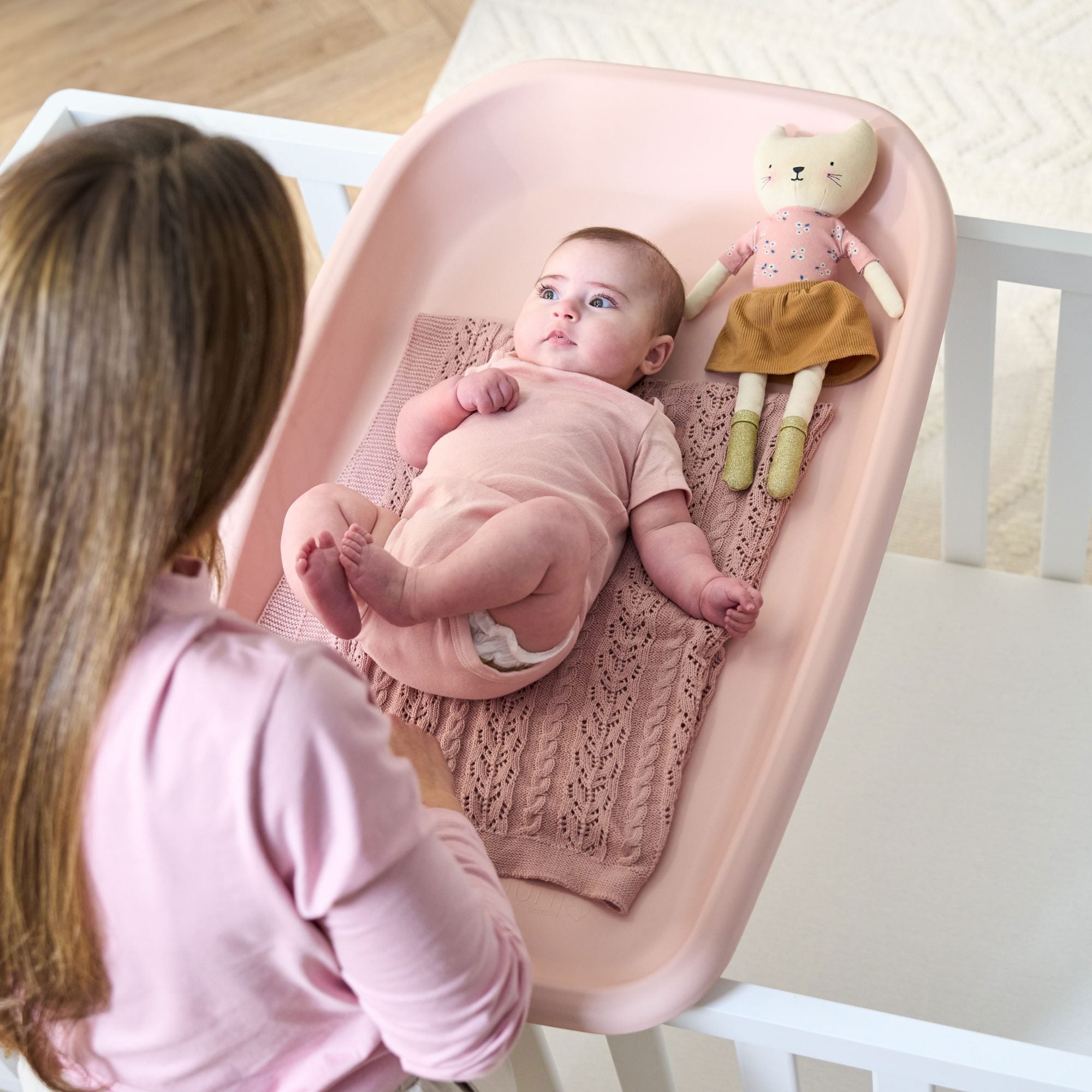Aerial view of mum changing baby on the Cushi mat in petal pink, showing contoured design, cosy blanket, and safety fit