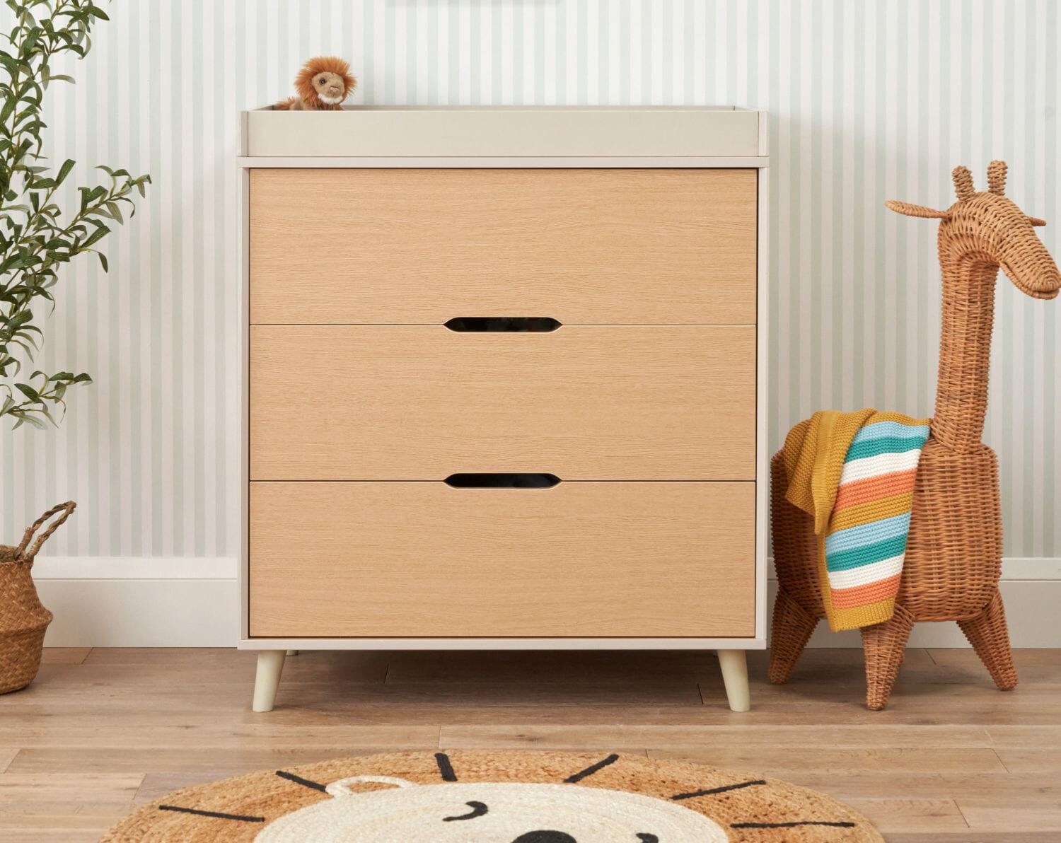 Front view of Fika chest of drawers in light oak with three drawers, splayed legs, and chic cut-out handles in a modern nursery with a giraffe toy.