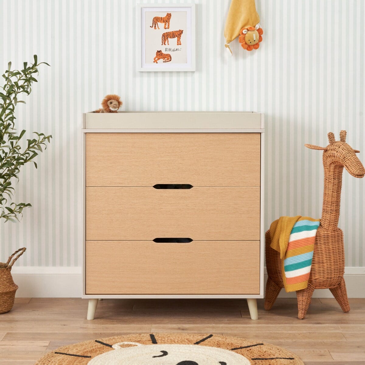 A Scandinavian-style chest changer with light oak and white sand colors, featuring stylish splayed legs and chic cut-out handles. The image also includes children's decorative items like a giraffe plush toy and a basket with a plant.