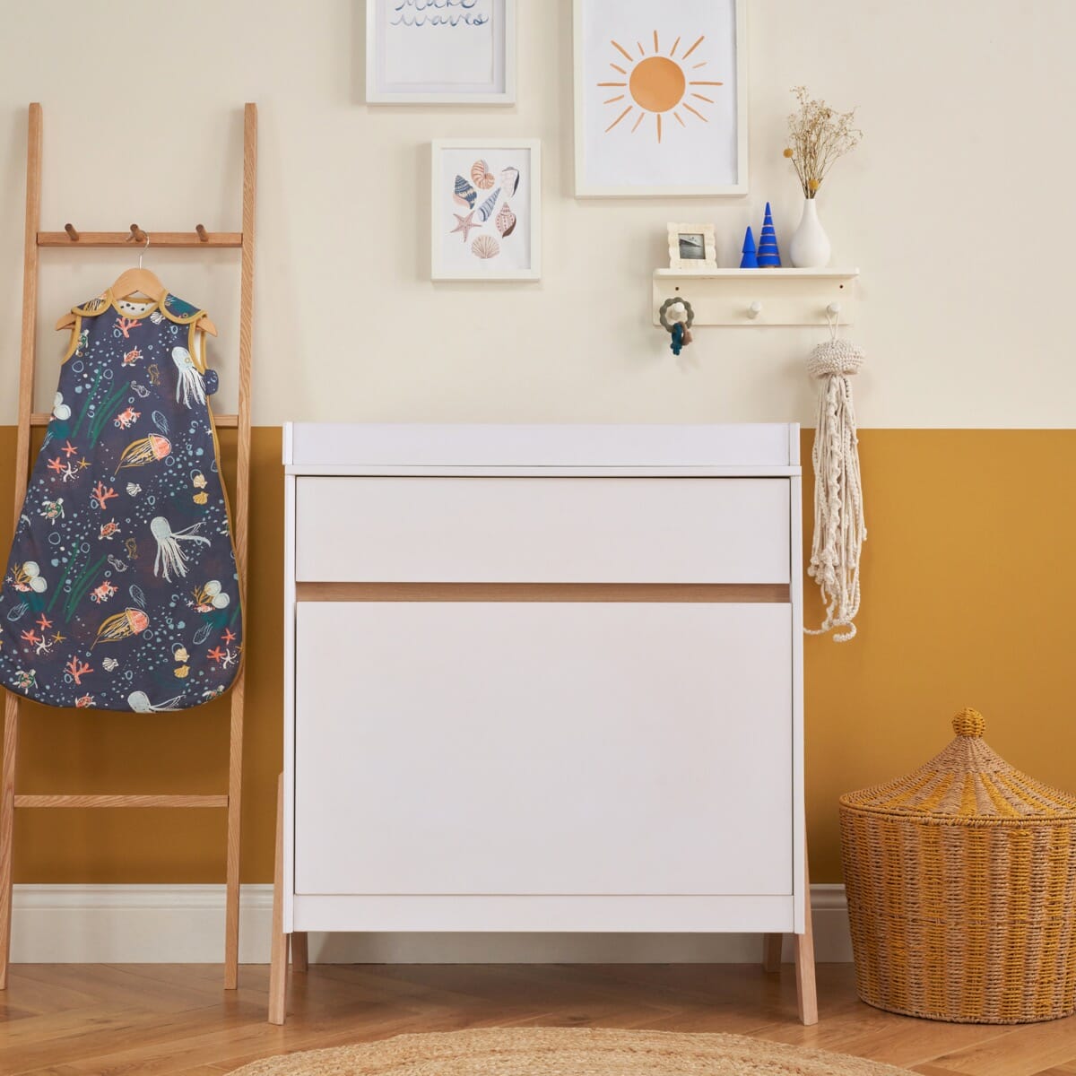 A white and light oak fuori chest changer with exposed legs and handleless drawers, situated in a room with a wooden ladder and a wicker basket