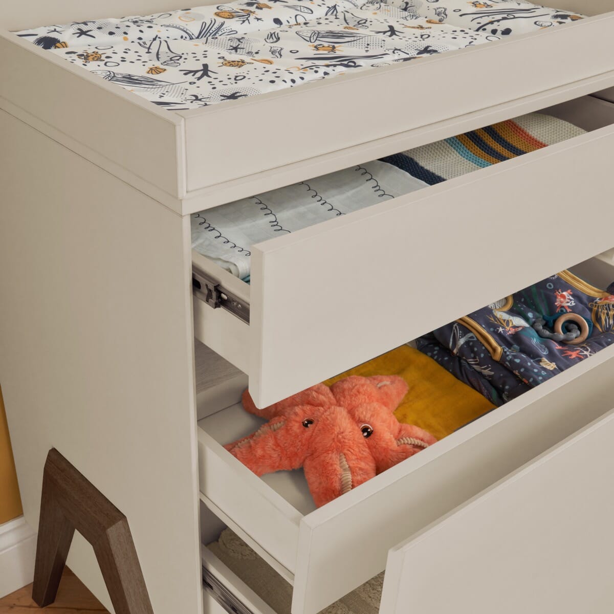 Angled view of the Fuori chest changer in white sand and warm walnut with smooth-glide drawers open, highlighting generous storage for baby essentials
